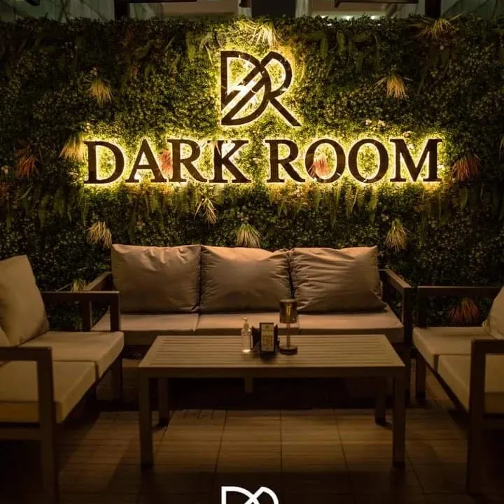 Illuminated Dark Room sign on a green plant wall above lounge seating